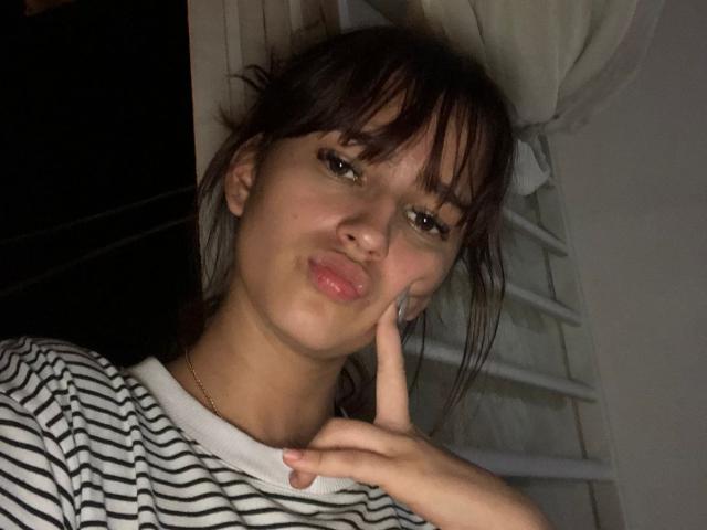 MelodyMansson has pouty lips, brown eyes, and bangs, wearing a striped shirt, with her finger on her cheek, looking at the camera, in dim light.