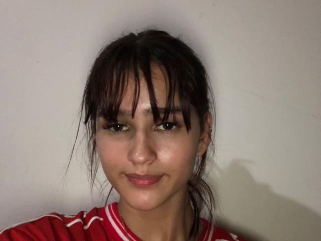 MelodyMansson with dark brown bangs, slight smile, wearing a red and white striped shirt, has light brown eyes, and a clear complexion, standing against a plain white wall.