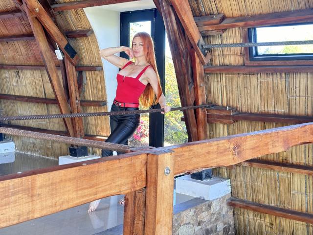 Leared with long red hair, red tank top, and black leather pants stands on a wooden bridge, leaning on a rope with one hand, showing her tits.
