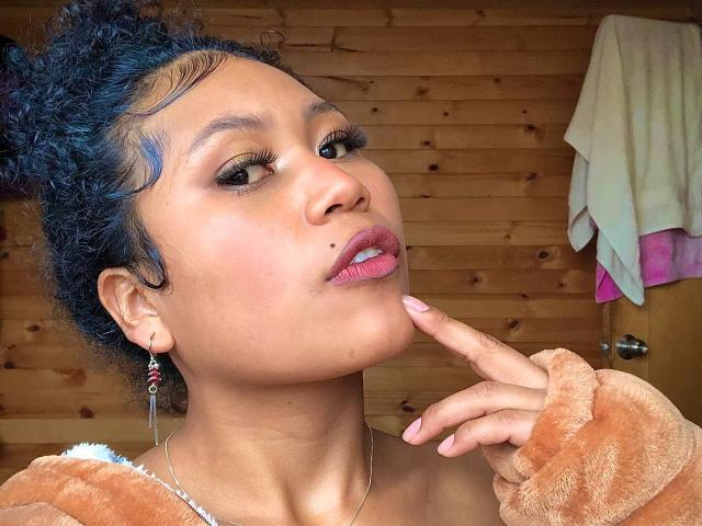 SophieSonyas has blue curly hair, brown eyes, pink lipstick, and a cute mole on her left cheek. She's touching her chin, wearing a fuzzy orange sweater.