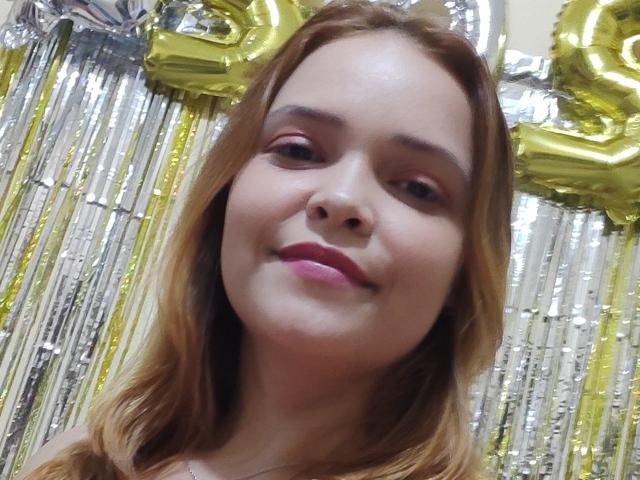 VickyMontero hat hellbraunes Haar, rosafarbenen Lippenstift und ein leichtes Lächeln. Ihr Gesicht ist zentriert, zeigt weiche Haut und braune Augen. Im Hintergrund befinden sich goldene und silberne Luftballons..