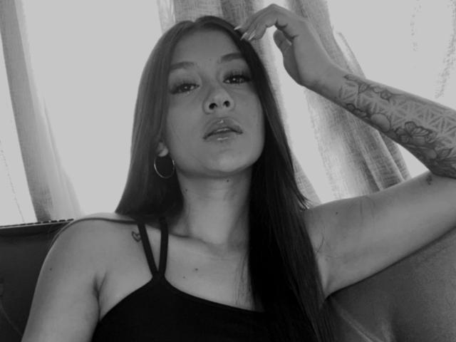 ThaliaAres with long dark hair, wearing a black tank top, has tattoos on her right arm, hoop earrings, and a relaxed expression, raising her right hand to her head.