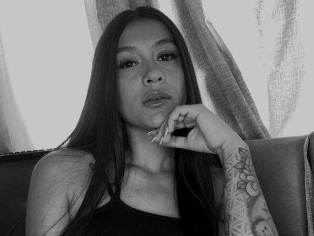 ThaliaAres has long dark hair, wears a black top, and rests her tattooed arm on a chair, looking at the camera with a slightly pouty expression in a black-and-white photo.