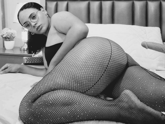 JackelineRoset con gli occhiali, sdraiata su un letto, indossa collant a rete e una maglietta nera, mostrando il sedere, guarda verso la telecamera, con un sex toy a destra..