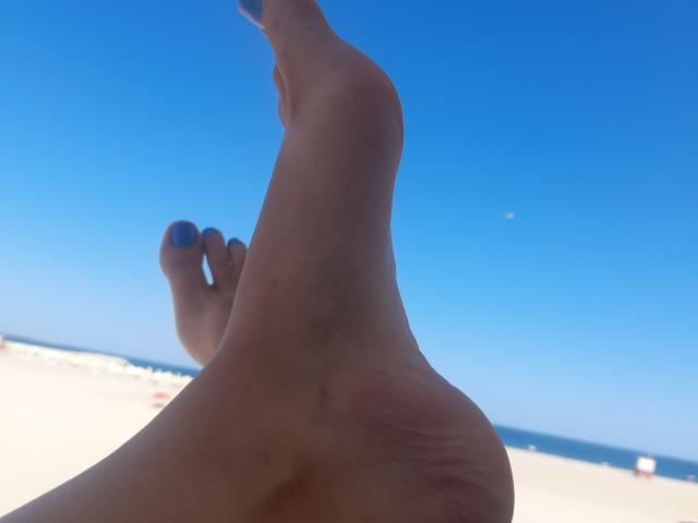 De blote voet van KateKate69, met blauw geverfde tenen, is in de lucht, waarbij de zool en de lichtrode hiel te zien zijn tegen een helderblauwe lucht..