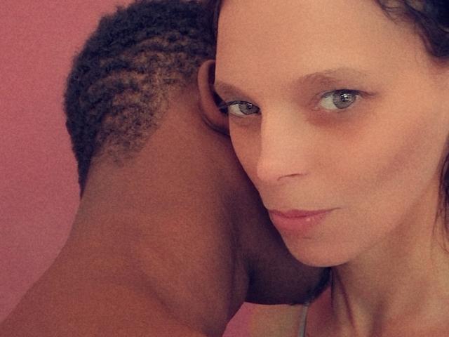 HotCouplez with dark skin and braided head, nose to nose with HotCouplez with light skin and blue eyes, both smiling slightly, pink background.