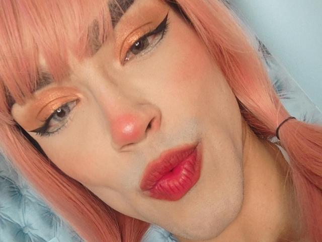 MelissaHotX69 tiene el pelo rosa, sombra de ojos naranja, delineador de ojos negro y labios rojos. Sus ojos están medio cerrados, lo que le da una expresión ligeramente fruncida. Su nariz es rosa y está mirando a la cámara..