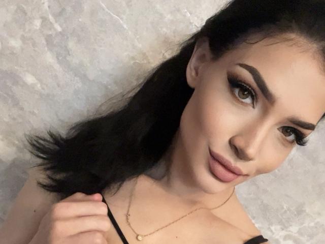 Moira Milicent mit schwarzen Haaren, dicken Augenbrauen und langen Wimpern, die ihr schwarzes Top mit Trägern hält, wobei ihr Brustansatz sichtbar ist, trägt eine goldene Halskette und lächelt in die Kamera..