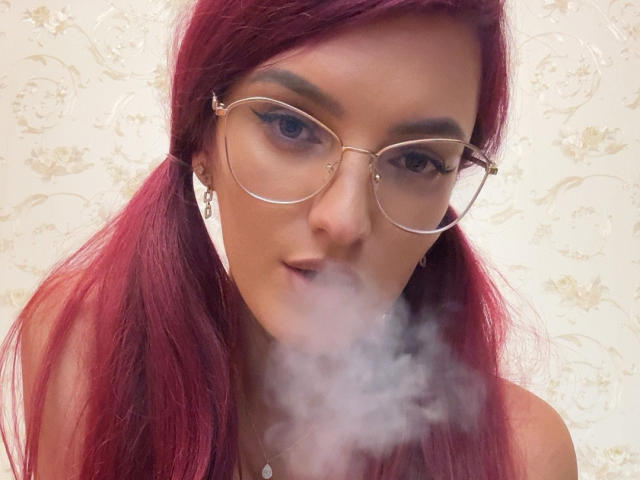 Rosalyn, con cabello rojo, una blusa de cuero negro y una liga de encaje, mostrando sus senos, sosteniendo un bolígrafo rosa, con uñas blancas y usando anillos, tomándose un selfie..