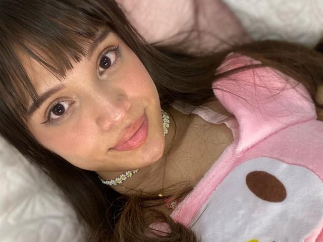 JackelineRoset acostada en una cama, con las nalgas levantadas, vistiendo una tanga negra, de espaldas a la cámara, con las piernas separadas, con un tapón anal visible, luz cálida en el dormitorio, las nalgas y la tanga de JackelineRoset en primer plano..