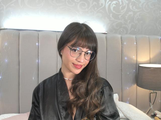 MelodySwet, com cabelos castanhos longos, óculos, um robe de seda preto e batom rosa, sorrindo para a câmera, sentada em uma cama com uma cabeceira cinza estofada e uma luminária..