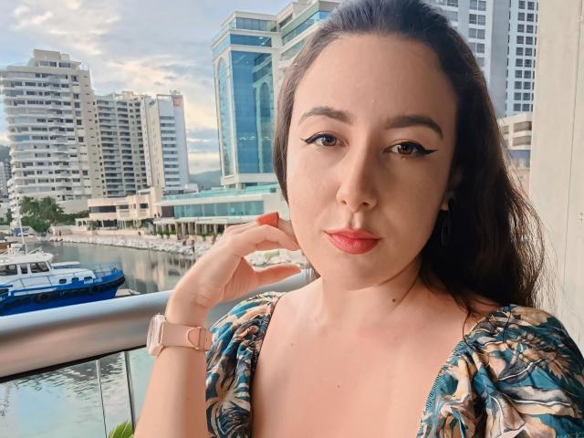 MabyLegrand with dark hair, wearing a floral dress, pink watch, and red lipstick, touching her chin, standing by a waterfront with tall buildings and boats in the background.