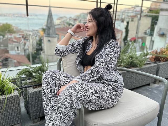 AmelieMuller, with long black hair in a bun, sits on a rooftop chair, wearing a zebra-patterned robe, exposing her black bra and showing her ass. She's smiling.