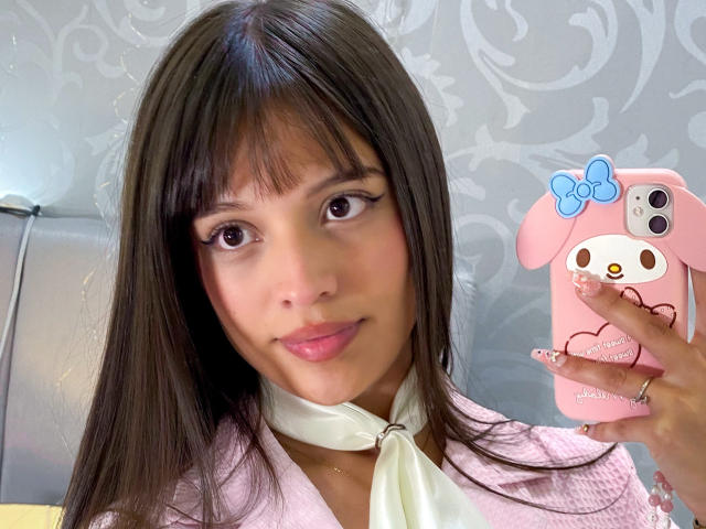 MelodySwet, con lunghi capelli castani, frangia e labbra rosa, tiene in mano un telefono rosa con una custodia a forma di coniglietto, indossa una maglietta rosa e una sciarpa bianca, e guarda verso la fotocamera..