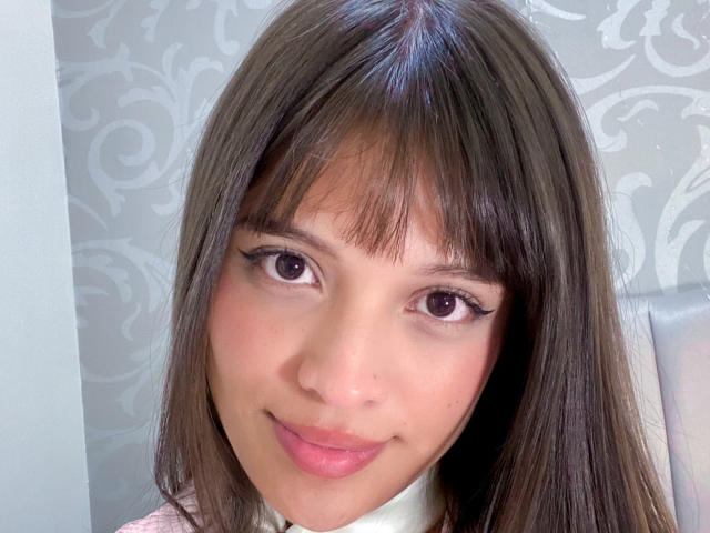 MelodySwet has brown eyes, straight brown hair with bangs, and a slight smile. She's wearing pink lipstick and a white shirt with a pink top. Her ass is not visible.