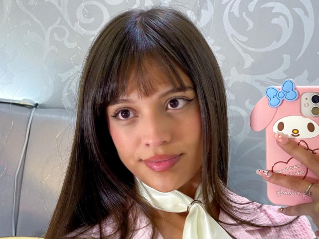 MelodySwet with long brown hair, bangs, and pink lips, holding a pink phone with a blue bow, wearing a pink jacket and white scarf, has cute eyeliner.