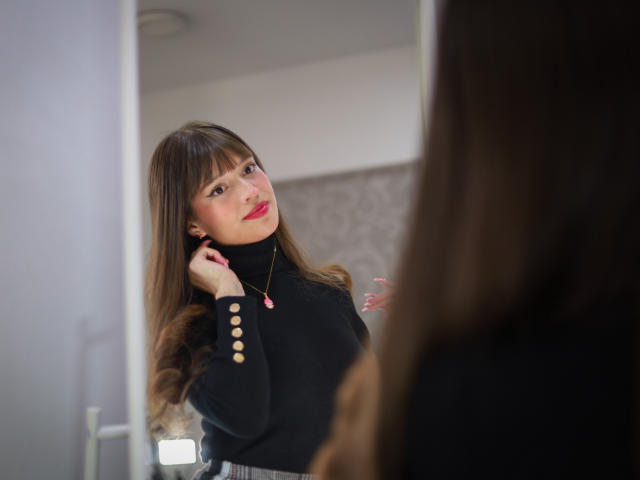 MelodySwet with long brown hair, black turtleneck, and gold necklace, looking at herself in a mirror, pink lips, gentle smile, right hand near ear, soft pink nails.