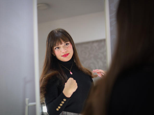 MelodySwet with long brown hair, wearing a black turtleneck, red lipstick, and a pink necklace, standing in front of a mirror, looking at her reflection with a slight smile.