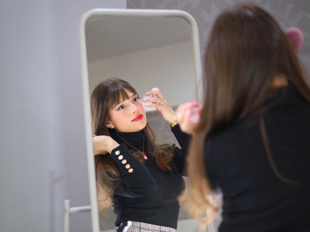 MelodySwet with brown hair, red lipstick, black turtleneck, and white skirt, applying pink eyeshadow in front of a mirror, showing her tits slightly in the reflection.