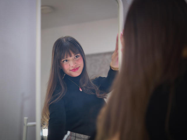 MelodySwet with long brown hair, black turtleneck, and plaid skirt, smiling at her reflection in a mirror, touching her cheek with a hand. Her tits are small.