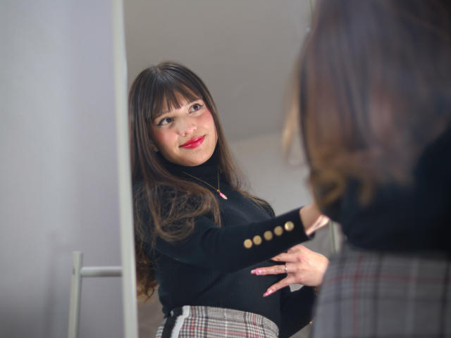 MelodySwet with long brown hair and red lipstick, black top, and plaid skirt, smiling at her reflection in a mirror, holding a black sleeve with gold buttons.