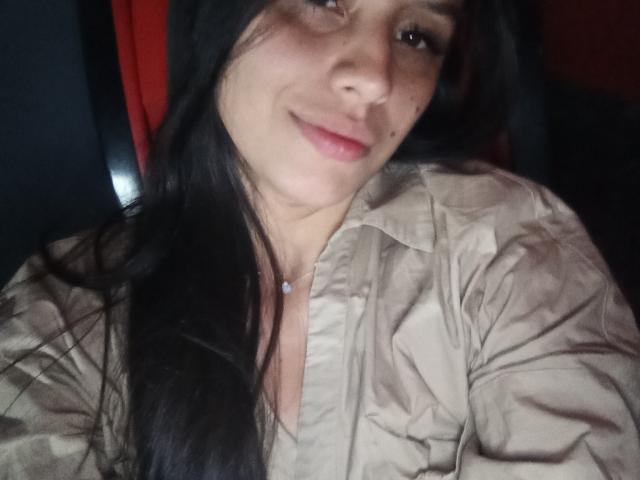 MadissonFontaine with long black hair, slight smile, light skin, wearing a beige shirt, small pink lip, necklace with white gem, medium-sized tits, dark background, slightly wavy hair.