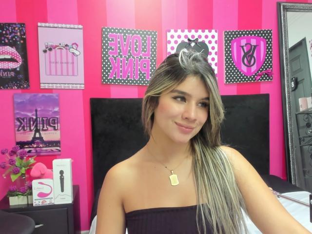 SashaPolet with long blonde and brown hair, wearing a strapless black top, smiling, pink and black room, black headboard, necklace with dog tag, black and pink wall decor.