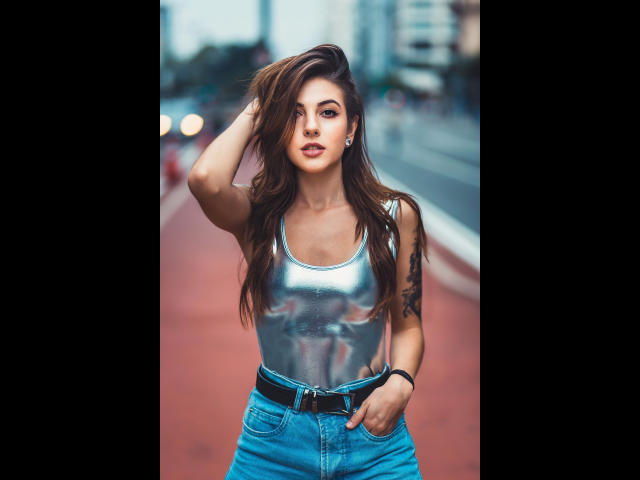 Consento-hot with long brown hair, wearing a shiny silver tank top and blue jeans, hand in pocket, black belt, tattoo on left arm, standing on a red track.