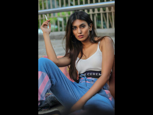 Consento-hot with long dark hair, wearing a white ribbed tank top and blue high-waisted pants, sitting with one leg up, holding a cigarette, looking at the camera.