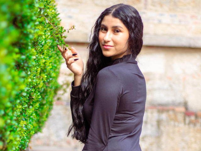 EmaHenderson with long black hair, wearing a black long-sleeve top, smiling, standing by a green bush, holding a leaf, looking over her shoulder.