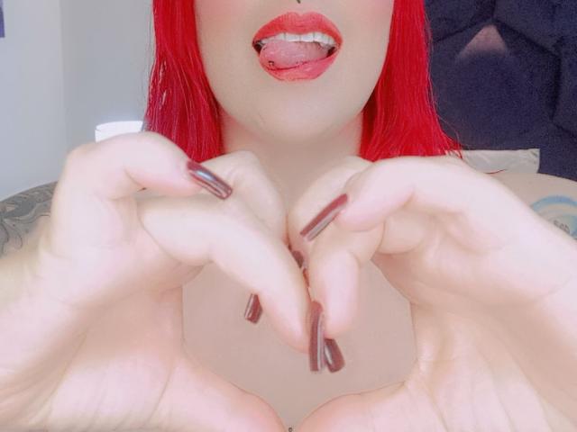 IsabellaRoshe with red hair, tongue out, showing a tongue piercing, holding her hands with dark red nails, tattoo visible on her right arm, and her boobs are slightly visible.