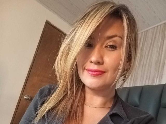 ValeryHottie69, con capelli biondi e castani, rossetto rosa, che sorride, indossa una maglietta nera, ha un viso carino e una sottile collana. Una porta sullo sfondo..