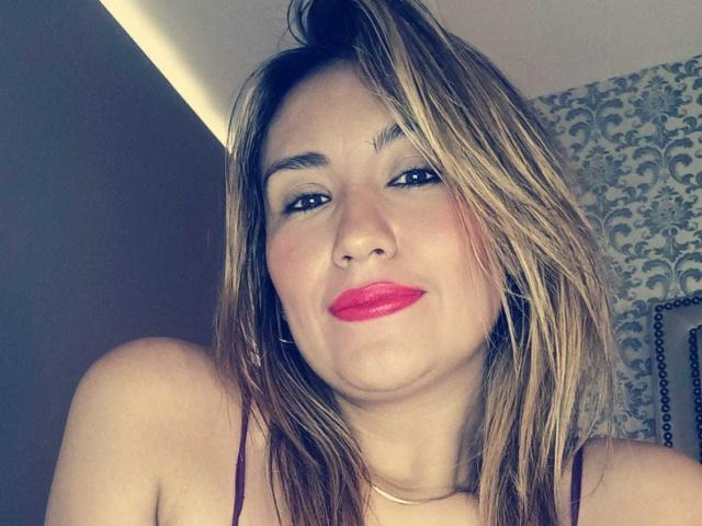 ValeryHottie69 tem cabelo loiro, batom rosa e está vestindo uma blusa vinho. Ela está olhando para a câmera com uma expressão levemente emburrada. Seus seios estão parcialmente visíveis..