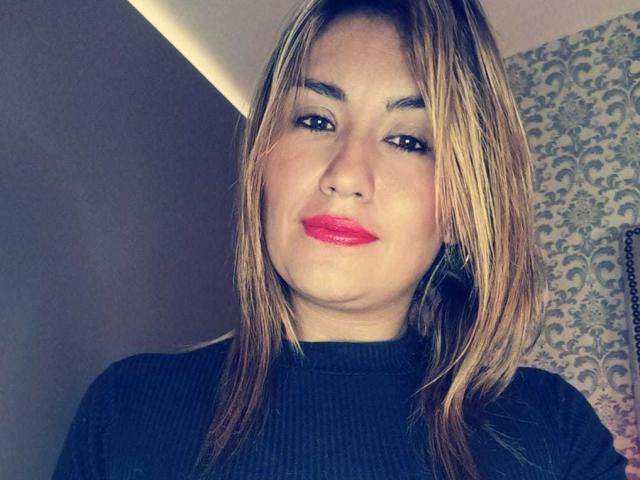 ValeryHottie69 heeft rode lippenstift, steil blond haar en draagt een zwarte coltrui. Ze glimlacht lichtjes met een schattig, mollig gezicht en bruine ogen..