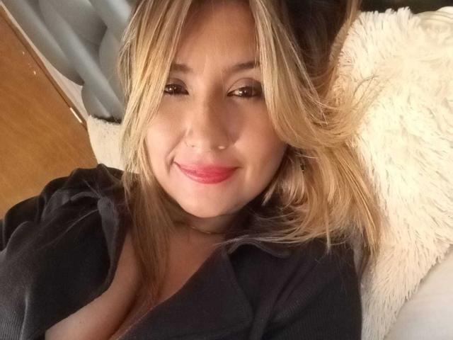 ValeryHottie69, con cabello rubio, lápiz labial rosa, blusa negra, un pequeño piercing en la nariz, sonriendo a la cámara, acostada sobre un almohadón blanco y esponjoso, mostrando un poco de escote..