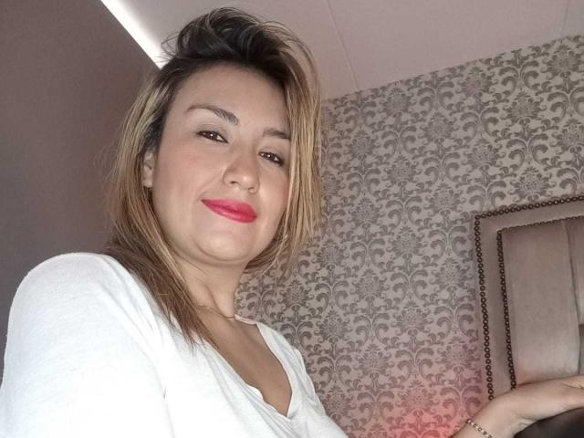 ValeryHottie69 heeft blond haar, roze lippenstift, draagt een witte blouse, heeft een licht opgetrokken neus en een zelfverzekerde glimlach, en staat in een goed verlichte kamer..
