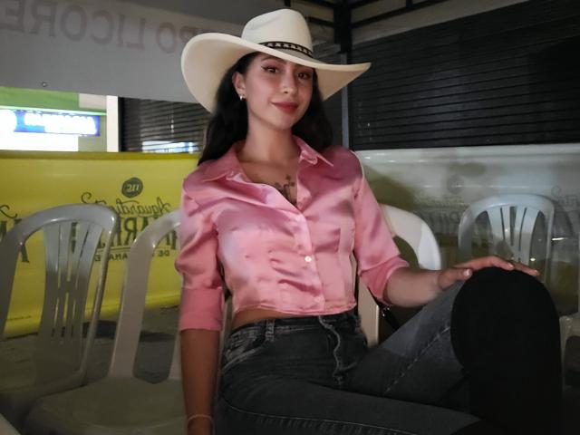 Emma Wells mit schwarzen Haaren, einem rosa Satinhemd, einem weißen Cowboyhut, blauen Jeans und schwarzen Leggings, sitzt auf weißen Stühlen, lächelt und zeigt ihre kleinen Brüste und ihr hübsches Hinterteil..