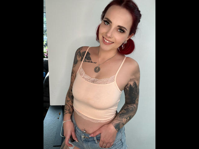NinaDevil, une femme aux cheveux roux, souriante, portant un débardeur rose avec des détails en dentelle, montrant des tatouages, et un jean bleu, debout avec les mains sur ses hanches, laissant entrevoir son nombril..