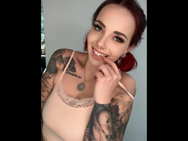 NinaDevil-hot with red hair, tattoos on arms and chest, smiling, pink lace bra, finger on lip, nose ring, earrings, ring, light skin, green eyes, and a cute smile.