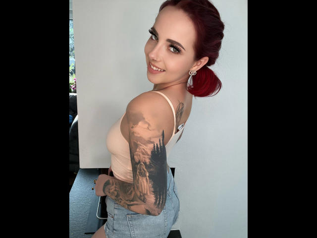 NinaDevil, une femme aux cheveux roux, souriante, portant un débardeur rose et un jean bleu, montrant un bras tatoué et un piercing au septum. Ses fesses sont visibles..