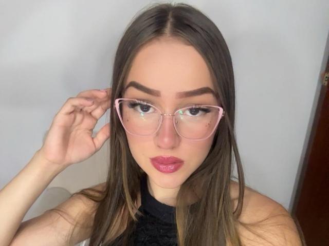 AntoniaSanchez has long brown hair, pink glasses, thick eyebrows, and glossy pink lips, with her hand near her face, wearing a black top, showing her tits slightly.
