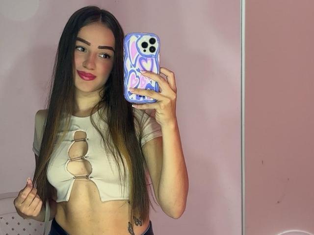AntoniaSanchez with long brown hair, red lipstick, wearing a cut-out white top revealing her small tits, holding a phone with a colorful heart pattern, taking a mirror selfie.