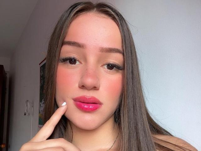 Antonia Sanchez, con lunghi capelli castani, labbra rosa e unghie bianche, che tocca la guancia con un dito, ha un viso carino e sopracciglia folte, e guarda la fotocamera..