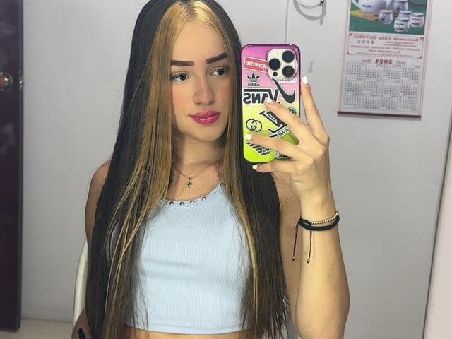 Antonia Sanchez, con lunghi capelli neri e biondi, rossetto rosa, canotta azzurra, che tiene in mano un telefono con una custodia colorata, selfie allo specchio, con il seno visibile, magra, sfondo bianco, trucco leggero..