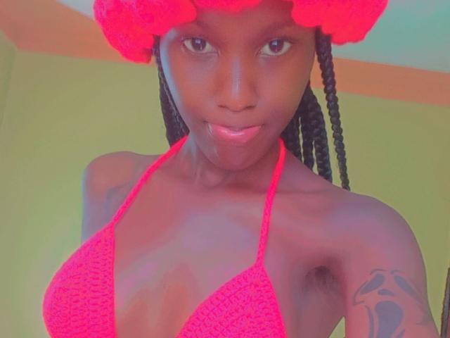 Sheila69s with dark skin, wearing a red crochet bikini top, red hat, and braids, showing small tits, armpit, and tattoo on her right arm. She's looking at the camera.