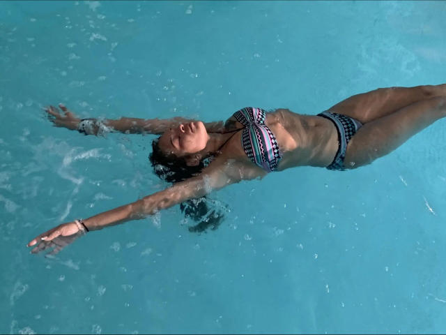 Brownsugarheaven-hot floating in blue water, arms out, wearing a patterned bikini, eyes closed, brown hair, tan skin, small tits, flat ass, water ripples around her.