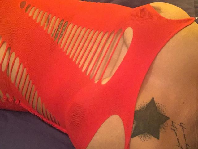 Brownsugarheaven-hot wearing a red, ripped-up, fishnet-like top, exposing her side and a green star tattoo on her lower back. Her skin is light, and she's lying down.