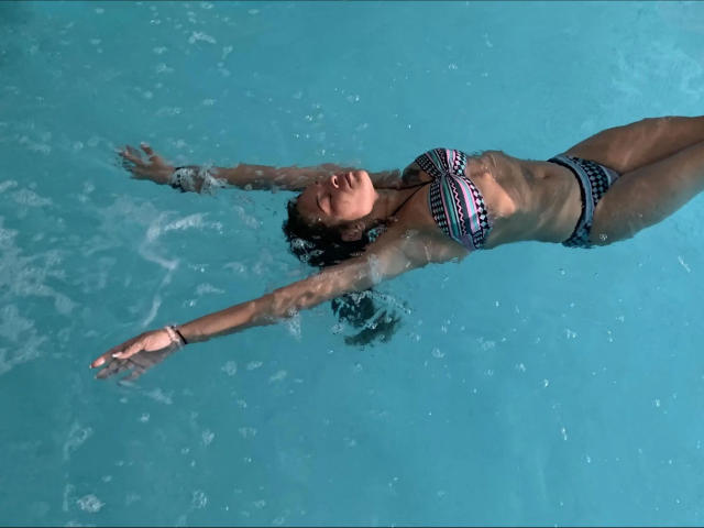 Brownsugarheaven-hot floats in a pool, arms out, wearing a colorful, patterned bikini. Her brown skin is visible, with water ripples around her. Her tits are small, and her pussy is not visible.