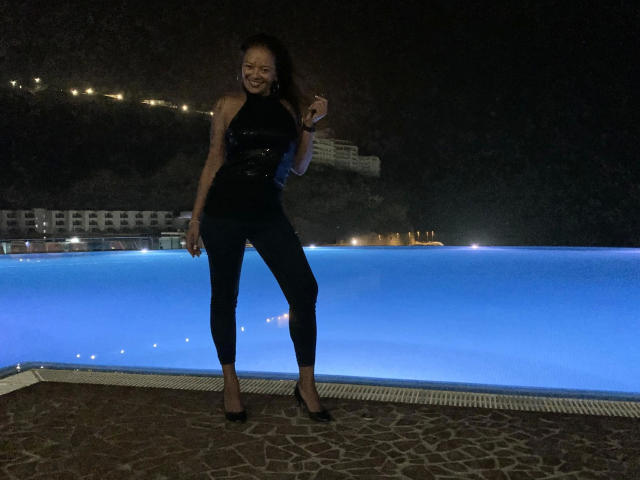 Brownsugarheaven-hot stands with a smile, wearing a black sleeveless top and black pants, blue-lit pool behind, night, dark background, long brown hair, black heels.
