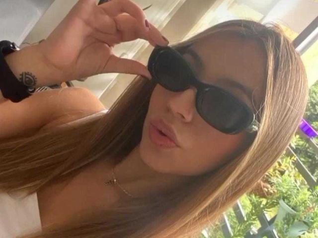 AmbarSkyler with long brown hair, wearing black sunglasses, pouting her lips, and touching her nose with her right hand, showing a small tattoo on her wrist.